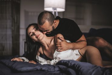 Ein heterosexuelles Paar möchte seinen Sex aufregender gestalten und verwöhnt sich halbnackt mit erotischem Vorspiel im Bett. Er liegt hinter ihr, nimmt sie sanft in die Arme und küsst ihren Hals.