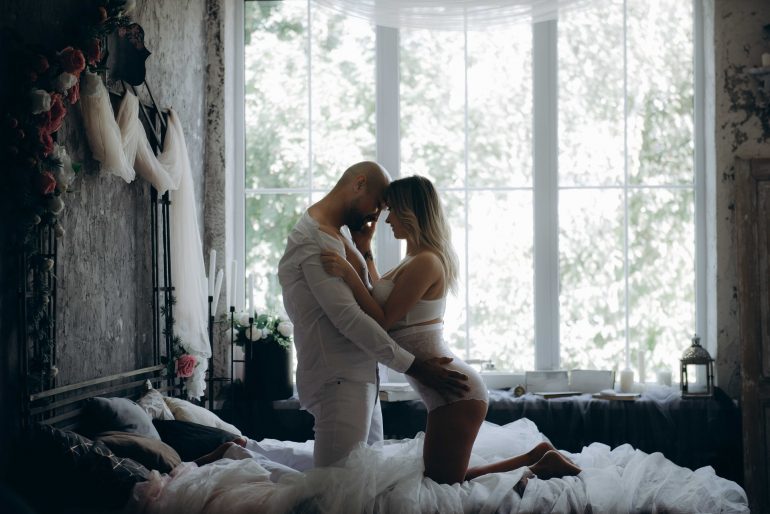 A newly married couple is romantically embracing on their marriage bed. She has taken off her wedding dress and is only wearing a negligee. Both look deep into each other's eyes and caress each other.