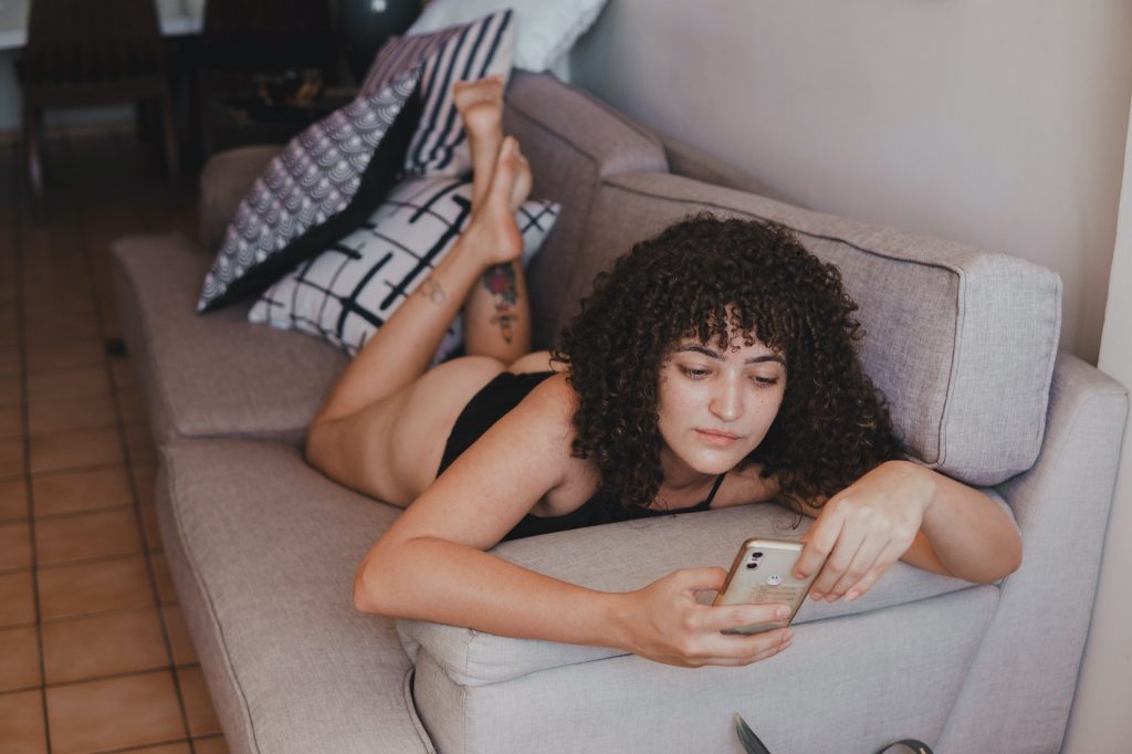 Eine Frau liegt halbnackt auf der Couch. Sie schaut interessiert auf ihr Handy und sucht mit Hilfe einer Dating-App nach sexueller Freiheit mit möglichen Partnern.