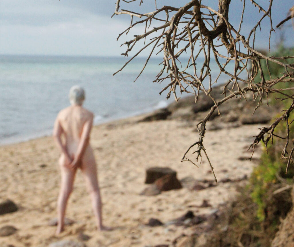Im unscharfen Hintergrund sieht man eine nackte, ältere Frau von hinten am Strand gehen mit Blick in die Ferne. Im Vordergrund sieht man die Äste eines Baumes.
