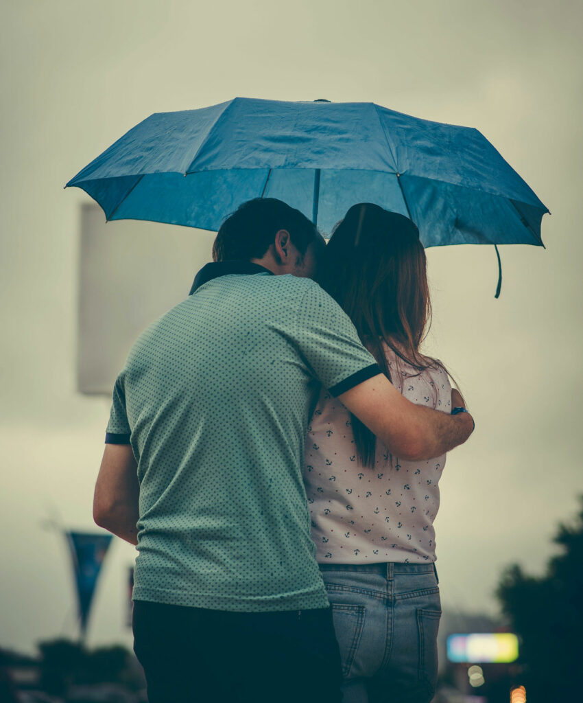 A couple stands together in the rain with a blue umbrella. The man holds the woman comforting in his arms.