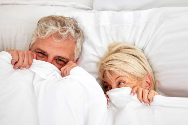 A middle-aged couple lies together in bed. Both are smiling, hiding their faces behind the white bedsheet.