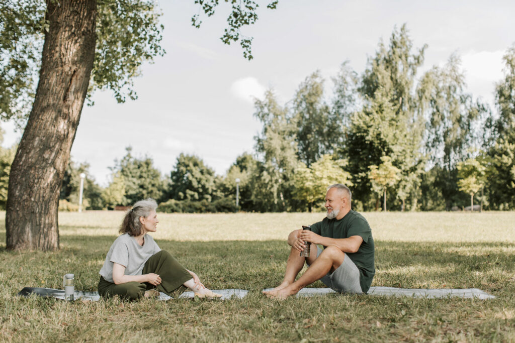 Two elderly individuals sit on separate mats in a park, smiling at each other and they're engaged in a conversation.