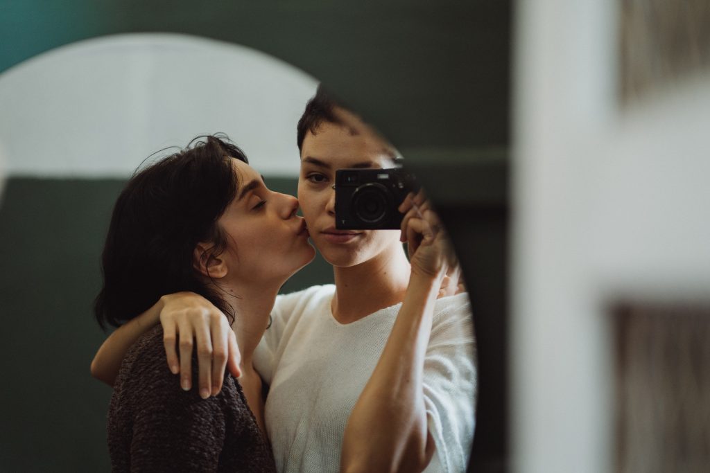A lesbian couple stands in front of a mirror holding a camera. They kiss and look forward to filming their exciting sex