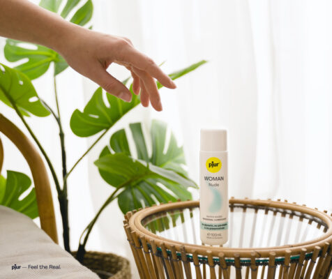 A snapshot of a pristine bedroom, featuring a bed, nightstand, plant, and white curtains. On the nightstand sits a bottle of pjur lubricant. A woman's hand reaches for it.