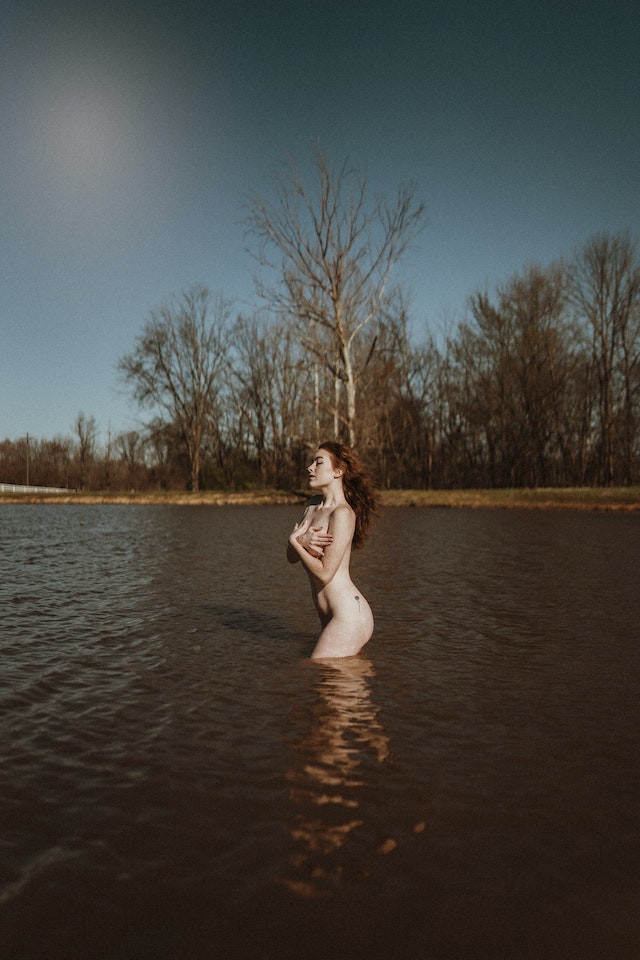 A naked woman is standing in a lake covering her breast with her hands