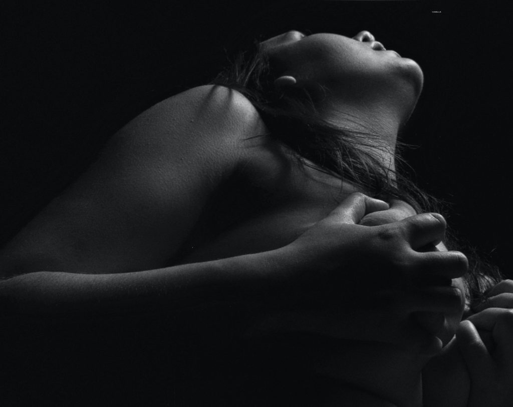 Ein Schwarz-weiß-Foto zeigt in Untersicht eine nackte Frau, die ihren Kopf in den Nacken gelegt hat und ihre Brüste mit ihren Händen bedeckt.