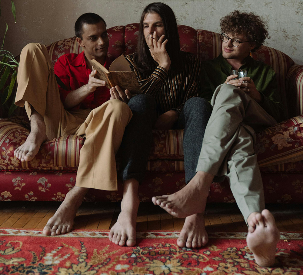 Three young men sit barefoot and relaxed on a colorful, older sofa, looking together into a book.