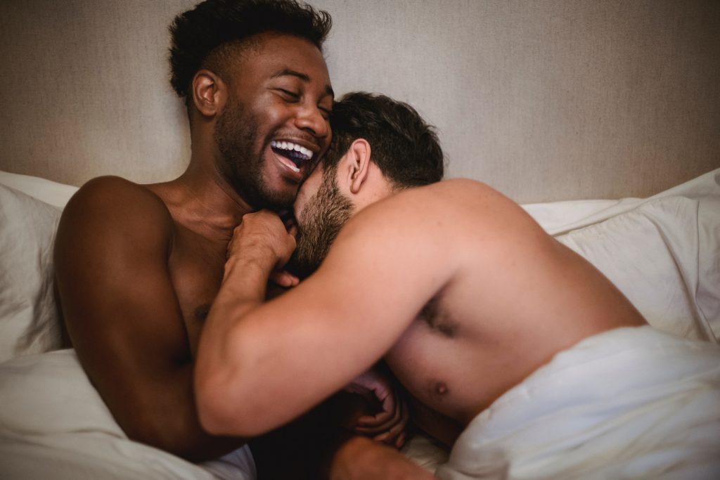 Two naked men are lying in bed and are hugging each other. They are laughing.