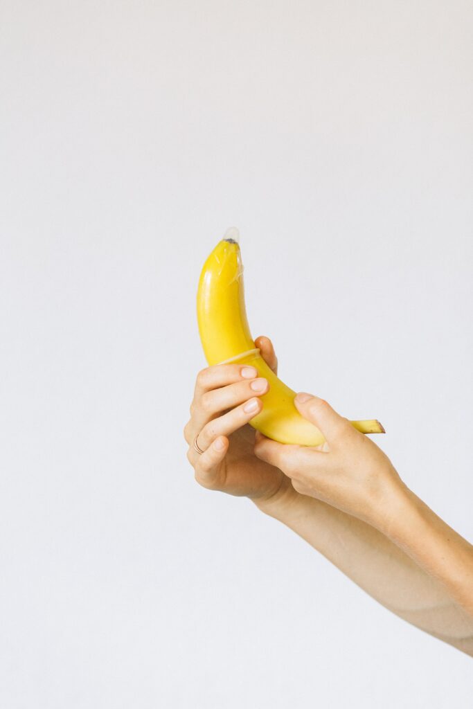 In focus is a hand holding a banana. Another hand pulls a condom over it. The background is white.