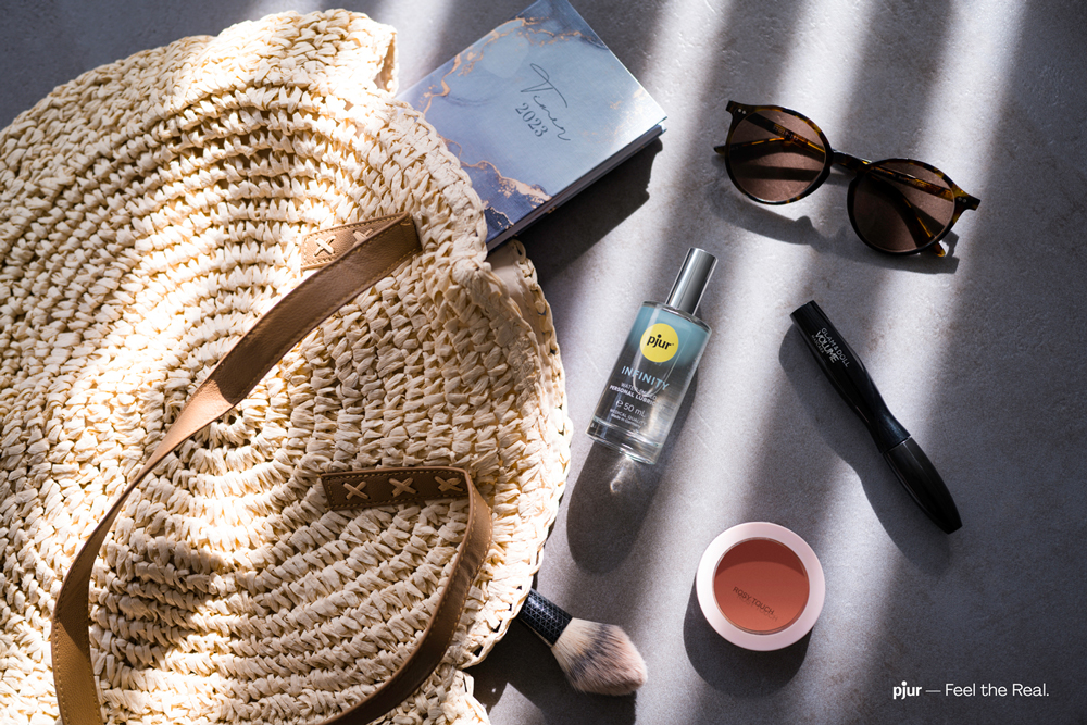 Blick von oben auf eine liegende Korbtasche, aus der verschiedene Gegenstände gefallen sind, zum Beispiel eine Sonnenbrille, Make-Up und eine Flasche von pjur INFINITY.