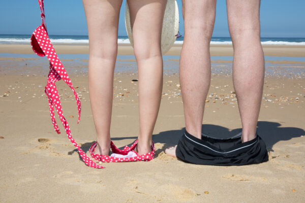 Man sieht die Beine eines Pärchens am Strand mit Blick auf das Meer. Beide Personen haben sich gerade ihrer Badekleidung entledigt.