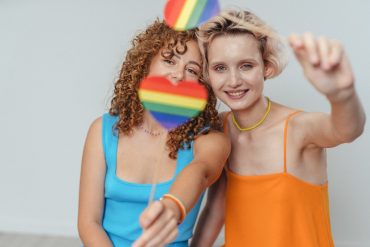 two women smiling and waving with the rainbow flag in a heart shape